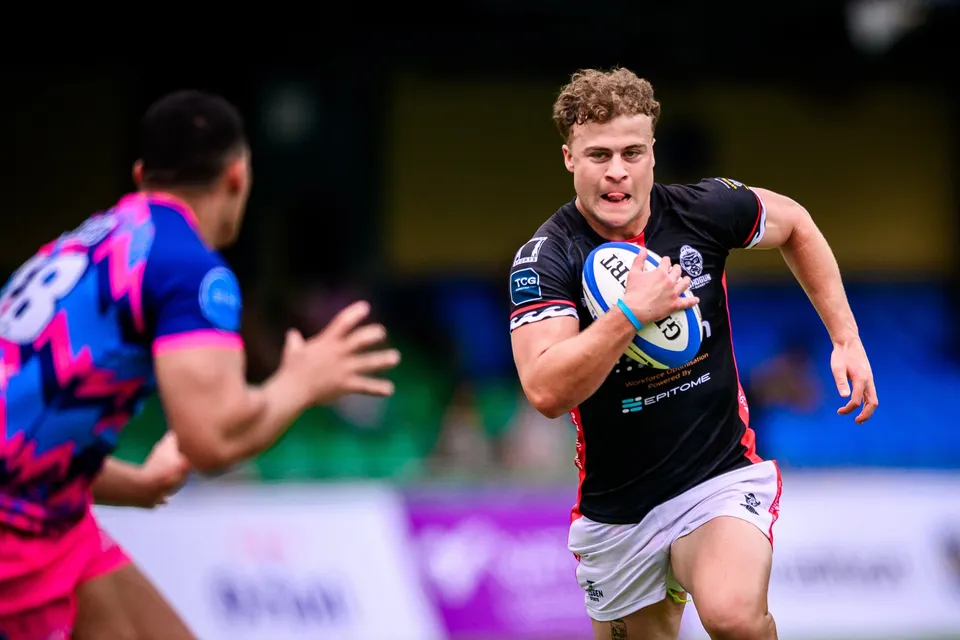 Curly-haired runner in black kit carries the ball at pace as a blurred OSOB defender reaches out in the foreground