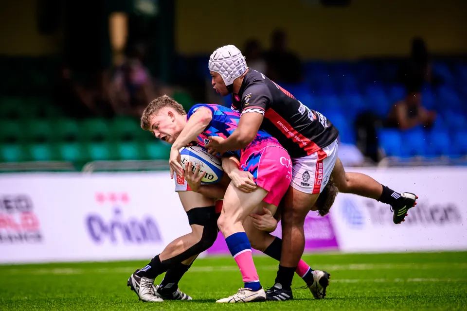 OSOB ball carrier is tackled by two black-shirted opponents, one in a white scrum cap, as he clutches the ball low