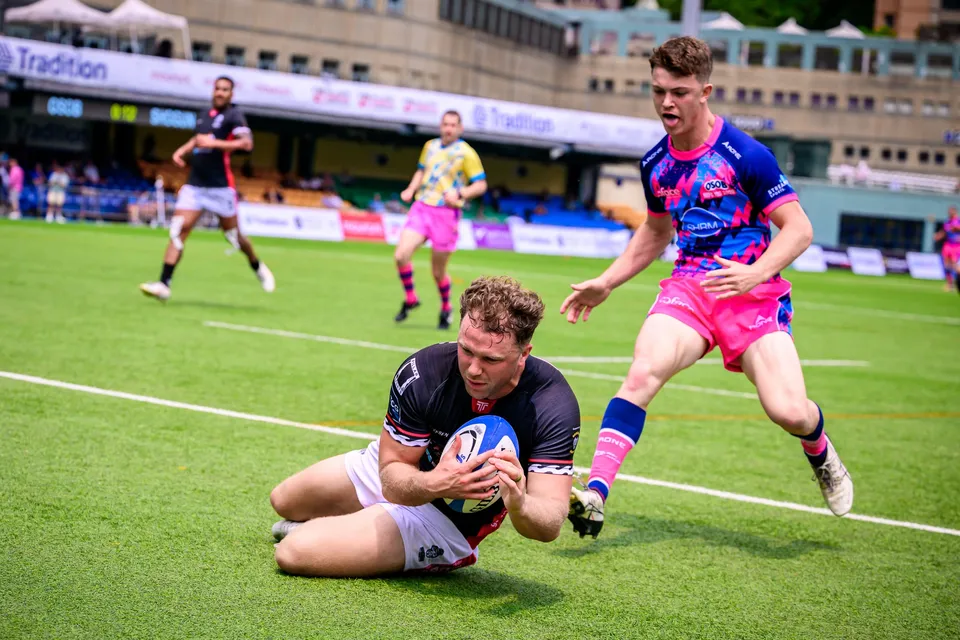 RGH Shogun player in black lies on the grass clutching the ball as an OSOB player in pink runs in behind