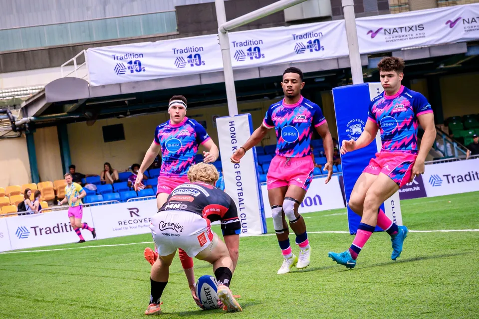 RGH Shogun player in black stoops over the ball as three OSOB players advance, Hong Kong Football Club boards behind