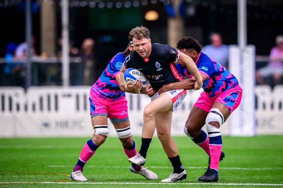Black-shirted Shogun ball carrier is gripped by two OSOB players in pink and blue patterned kit, 2026 match
