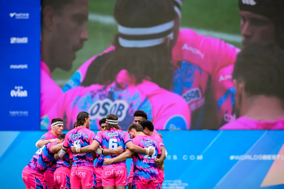 OSOB players in pink and blue shirts huddle with arms linked on the pitch in front of a big screen