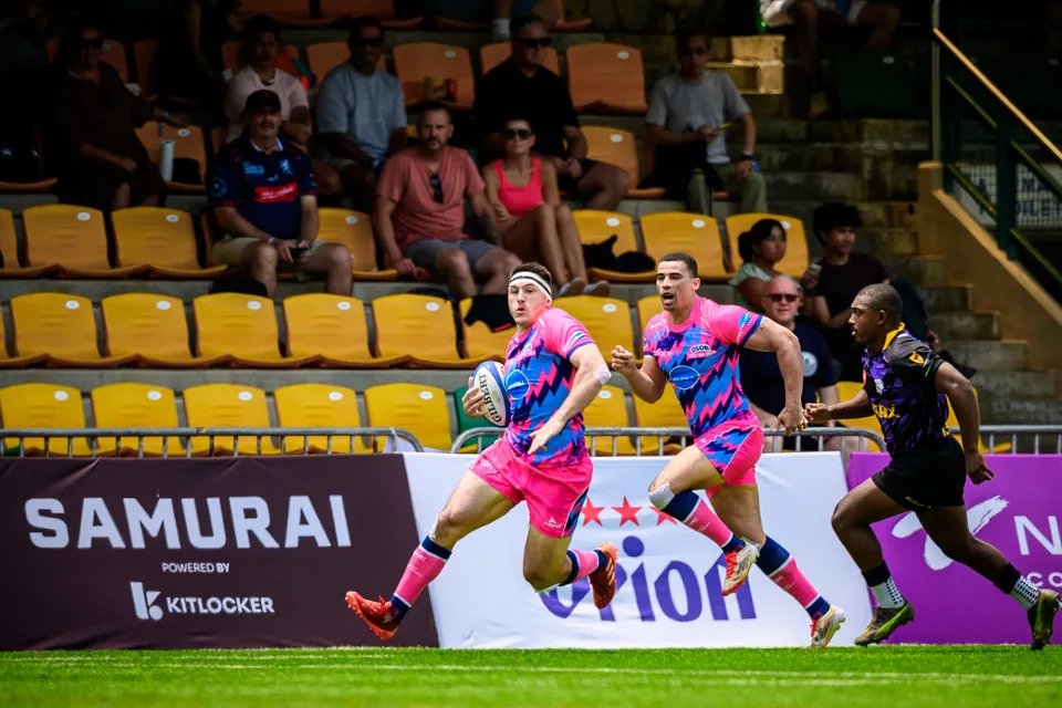 Two OSOB players in pink run in support with the ball past a Samurai banner as a purple defender chases