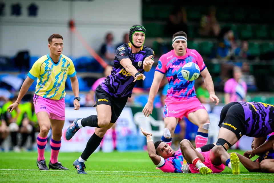 Headband-wearing OSOB player holds the ball while a purple-kit defender reaches out and a team-mate lies on the grass