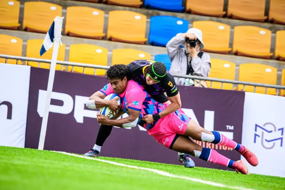 OSOB player in pink dives for the corner with the ball as a purple-kit defender tackles him beside the flag