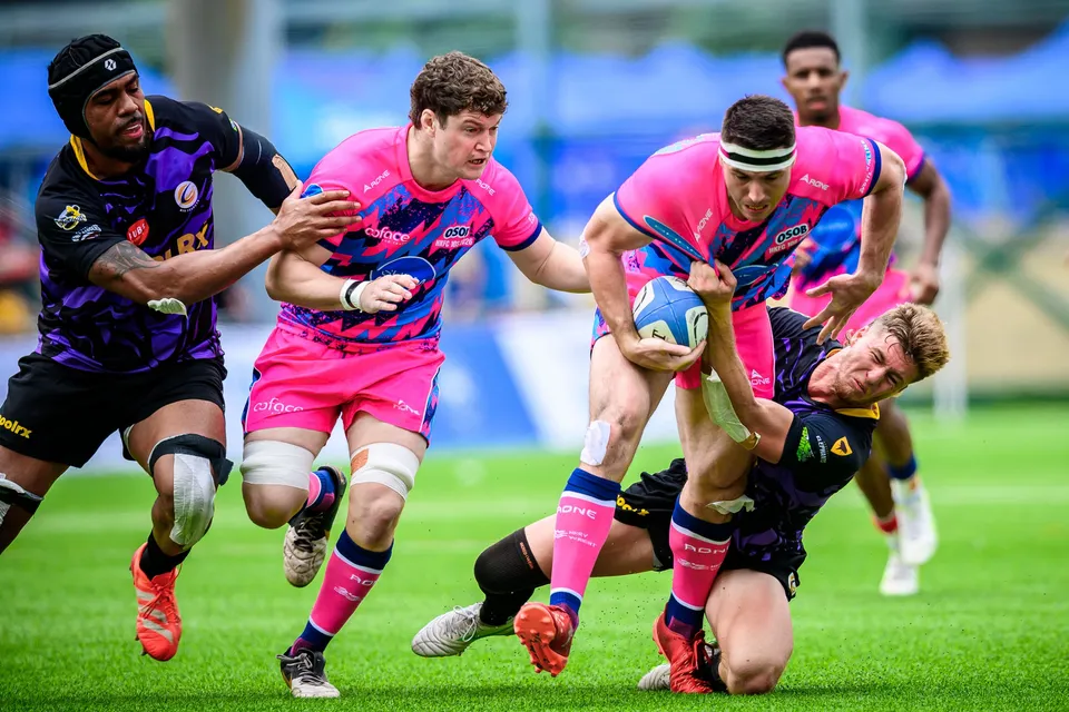 OSOB ball carrier is tackled low by a Rugby Tens Selects defender as pink-shirted team-mates run up in support