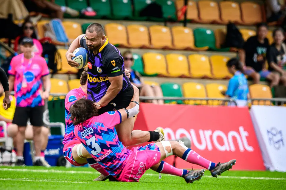Purple-kit ball carrier is tackled low by an OSOB number 3 in front of a red hoarding and a yellow stand