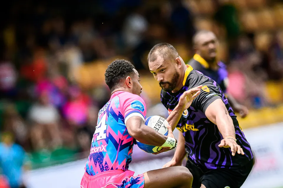 OSOB ball carrier fends off a bearded Rugby Tens Selects player in a close-up with the crowd blurred behind