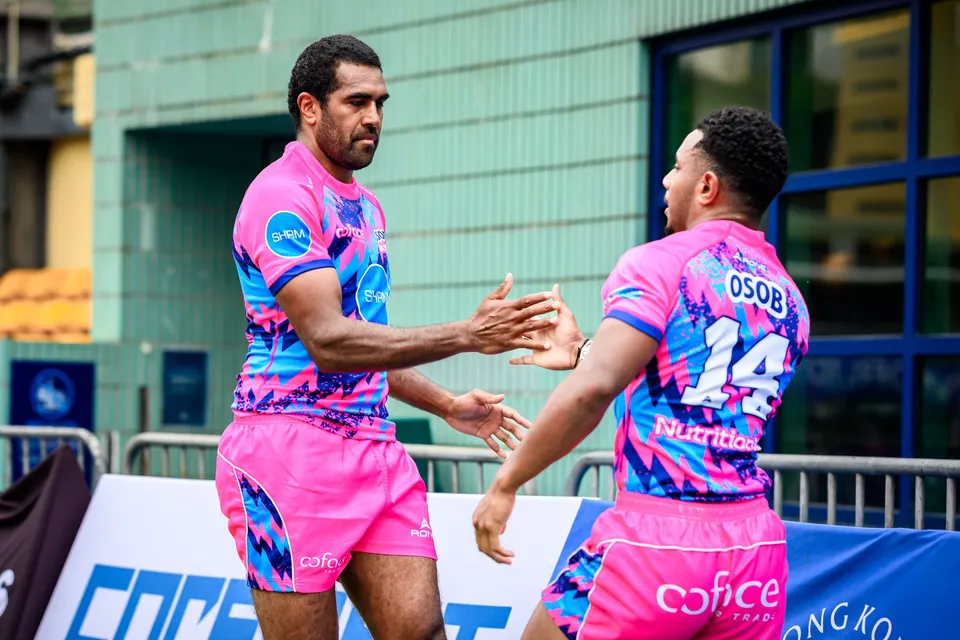 Two OSOB players in pink and blue clasp hands beside the touchline boards, one wearing number 14
