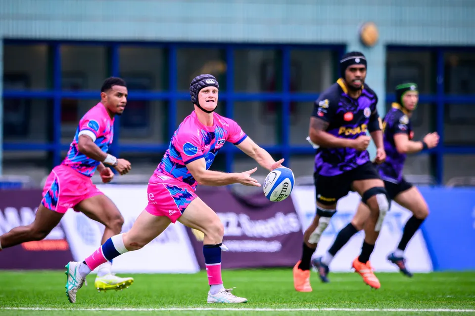 OSOB player in a scrum cap passes the ball with both hands as a team-mate runs beside him and purple defenders follow