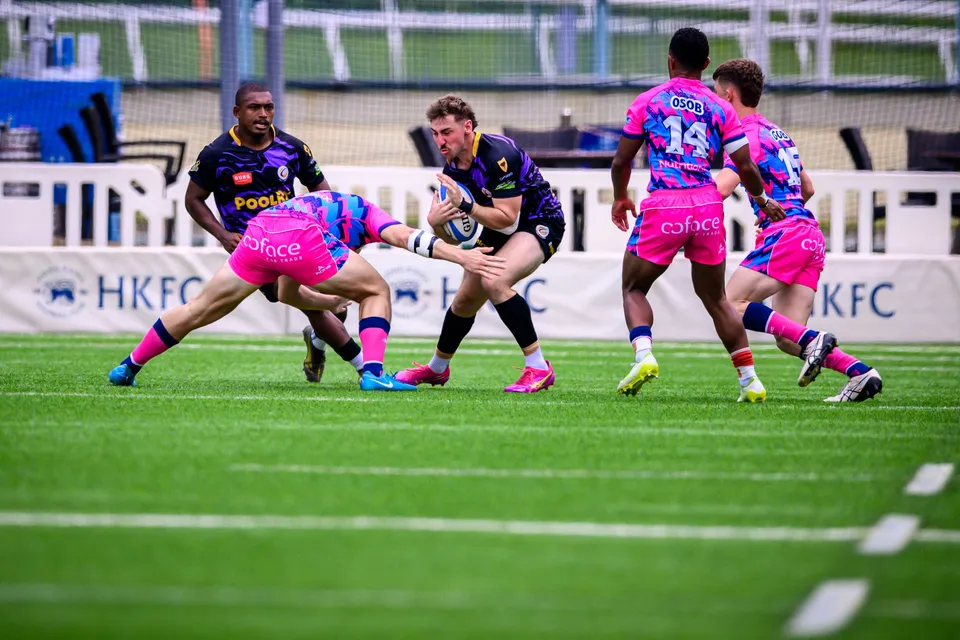 OSOB player ducks low into a tackle on a purple-kit ball carrier along the HKFC boards, number 14 close by