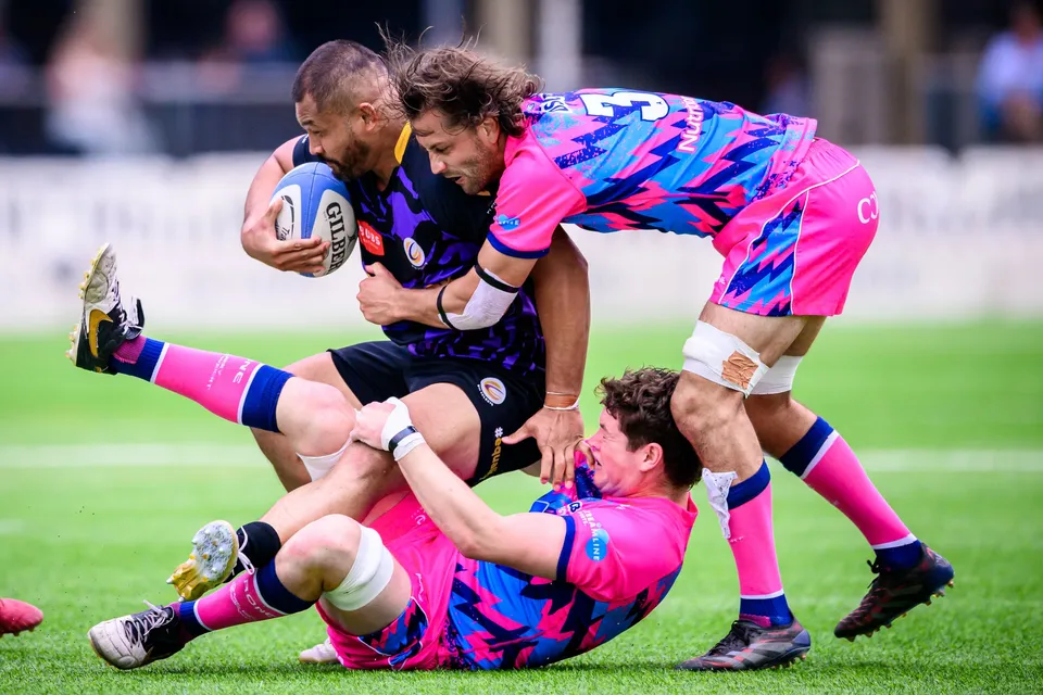 Purple-kit ball carrier is wrapped up by a standing OSOB tackler while a second OSOB player lies on the grass beneath
