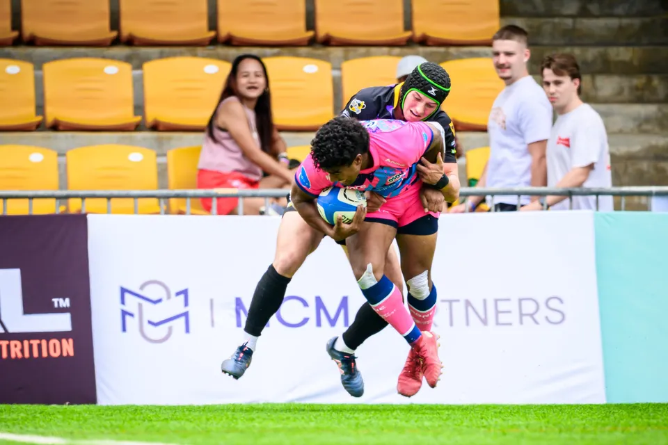 OSOB ball carrier is lifted off the ground by a Tens Selects defender in a green headguard beside the advertising boards