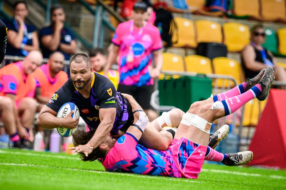 Tens Selects ball carrier in purple is brought down on the grass by an OSOB player, spectators in the stand behind