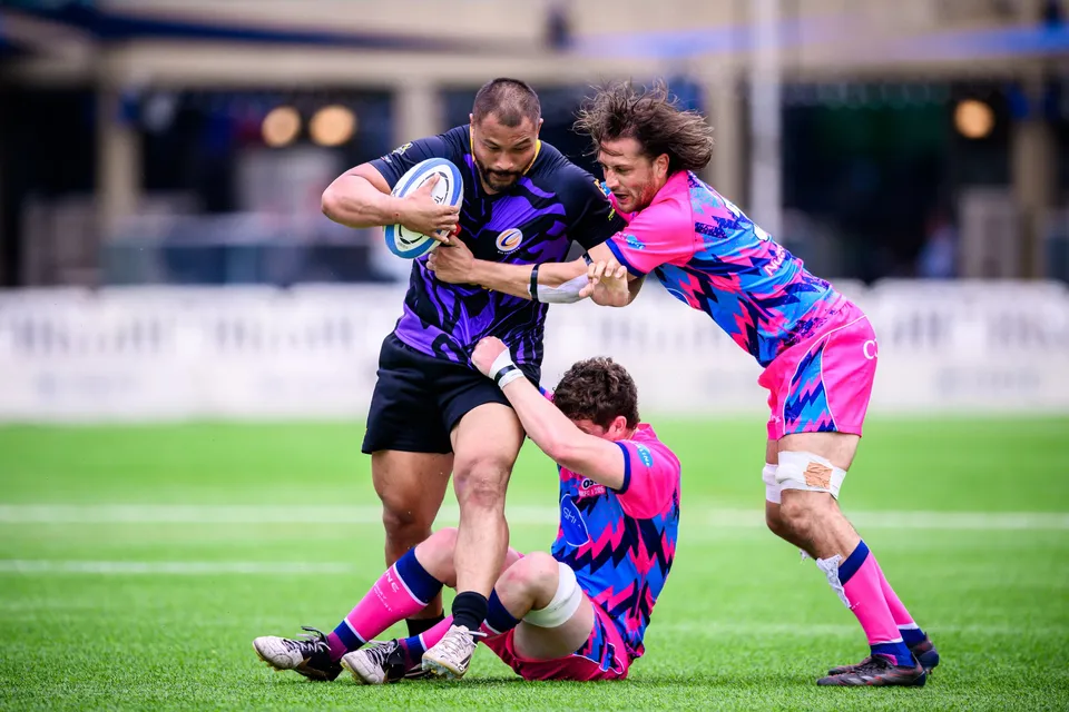 Large Tens Selects ball carrier is held by a long-haired OSOB player while a teammate sits on the grass