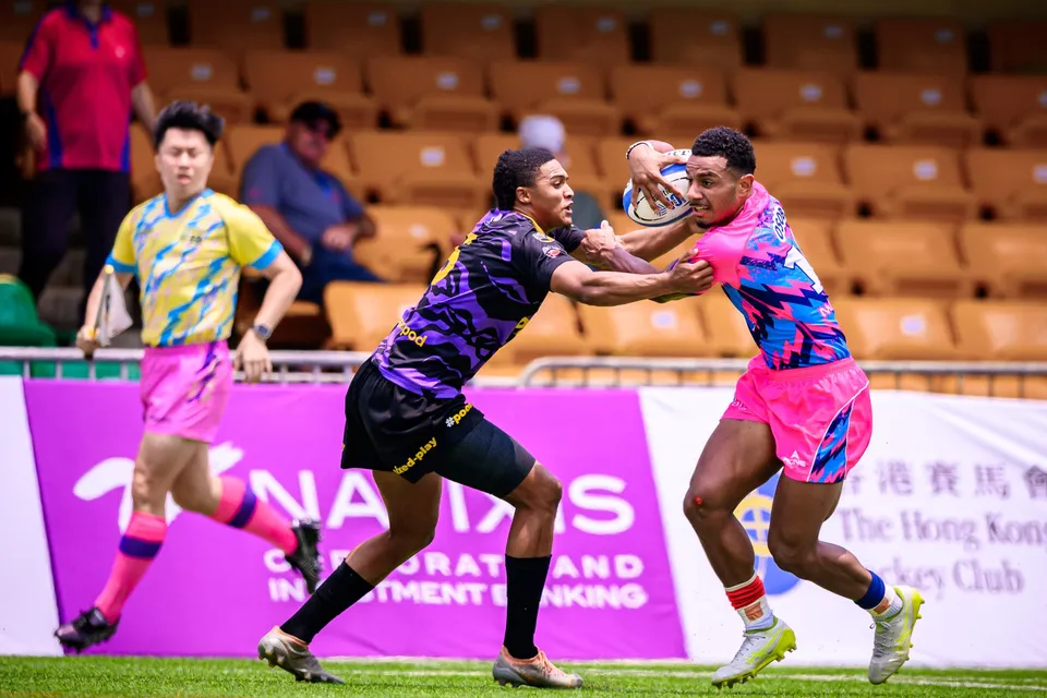 OSOB player fends off a Tens Selects defender with one arm while carrying the ball, an official and banners behind