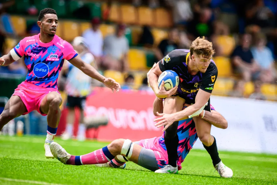 Rugby Tens Selects ball carrier in purple is dragged down at the ankles by a sprawling OSOB defender in pink