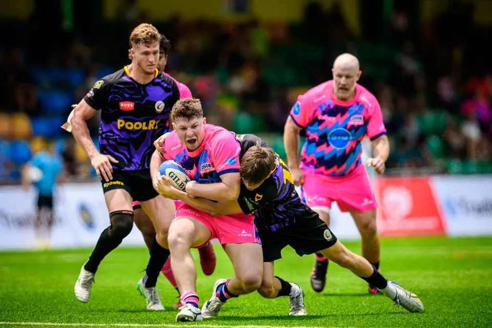 OSOB ball carrier in pink is gripped by two purple-kit Rugby Tens Selects tacklers as a team-mate runs up