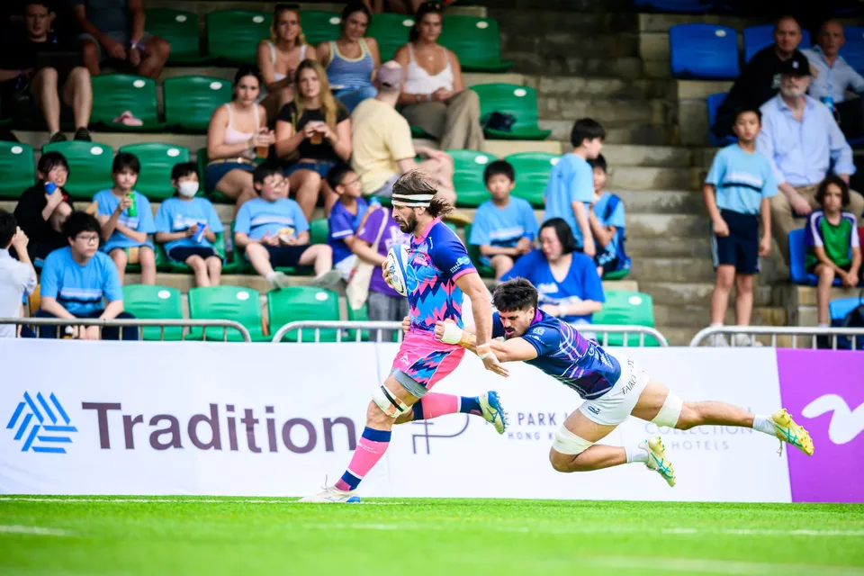 OSOB runner in a headband evades a diving Exiles tackler along the touchline as children watch from the stand