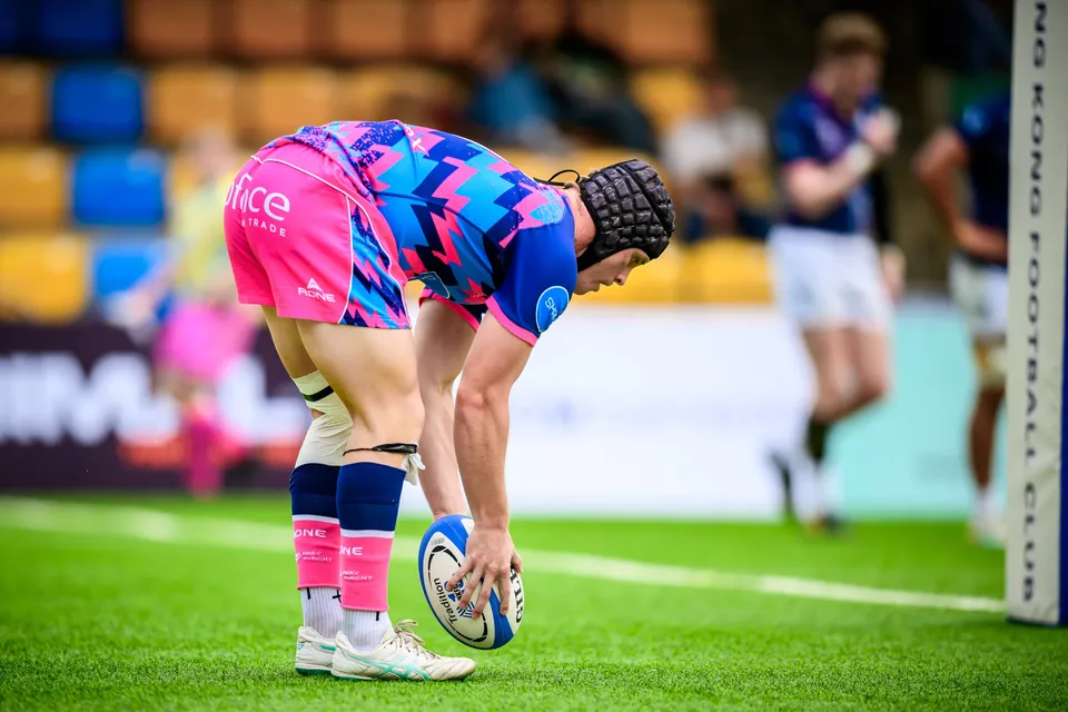 OSOB player in a scrum cap bends to place the ball on the grass beside a Hong Kong Football Club post
