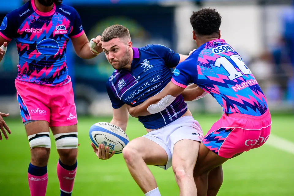 Scottish Exiles ball carrier in navy is wrapped up by an OSOB defender in pink, with a teammate looking on