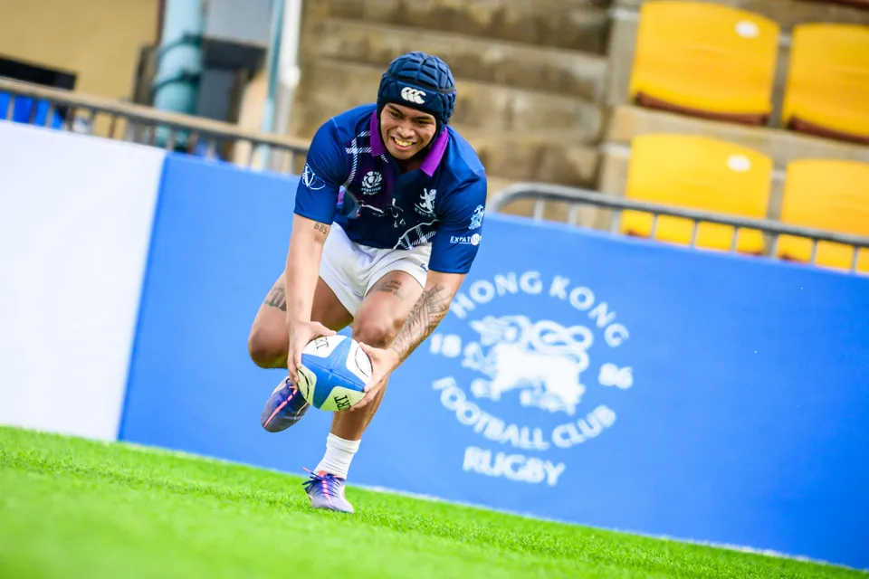 Exiles player in a navy skull cap grins as he lunges forward with the ball in front of a blue HKFC board