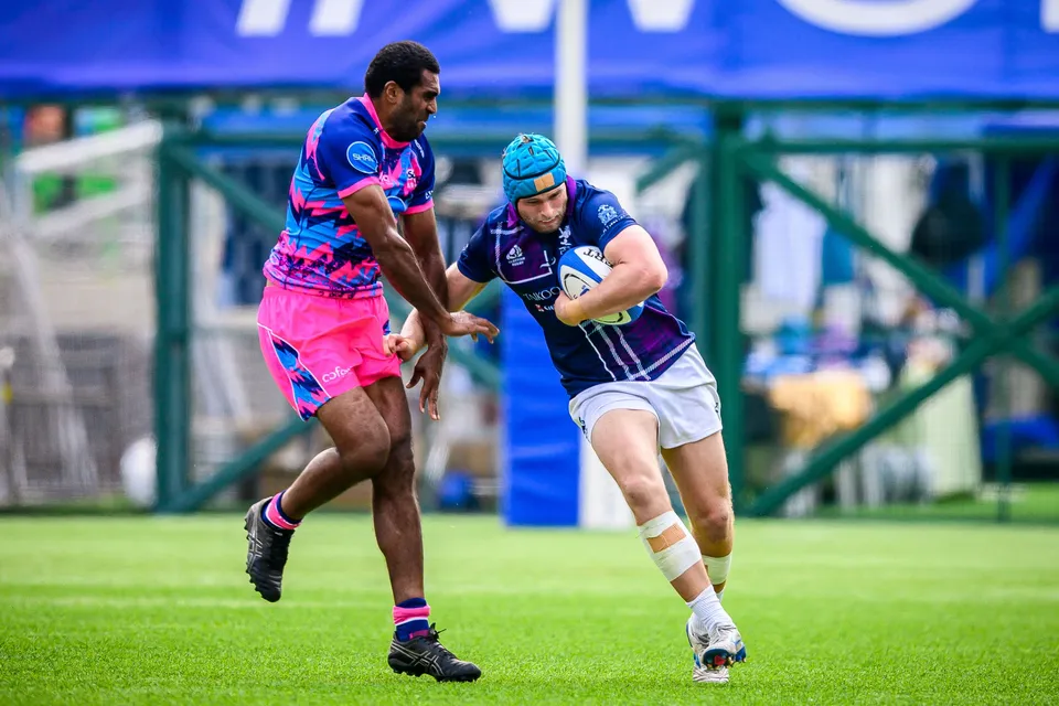 Scottish Exiles runner in a turquoise skull cap carries the ball past an OSOB defender in pink with arms out
