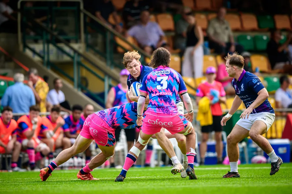OSOB number 3 in pink carries the ball into contact as an Exiles player waits, substitutes behind the barrier