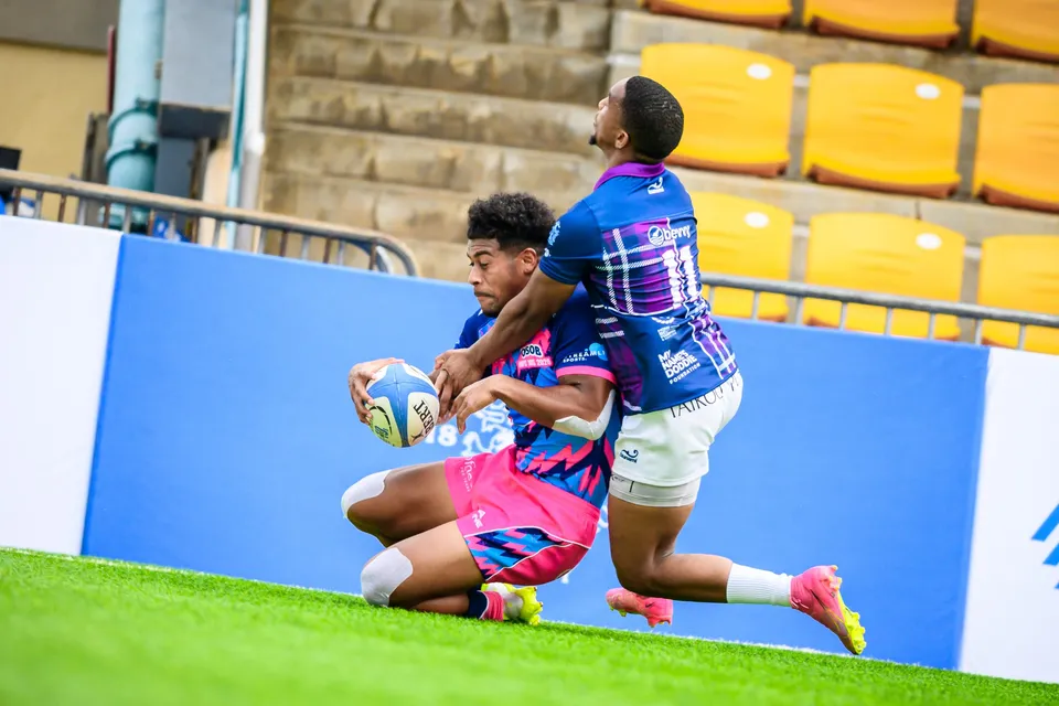 OSOB player in pink slides on his knees holding the ball as an Exiles player leans over him by the blue board