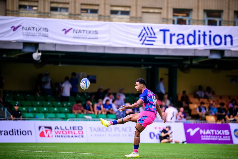 OSOB player in pink boots the ball high in front of Natixis and Tradition banners and a seated crowd