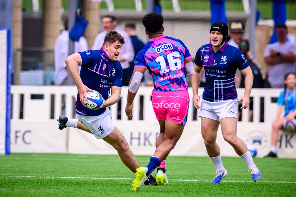 Exiles ball carrier in navy runs at the camera as OSOB number 16 in pink looks on and a teammate follows