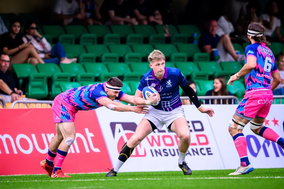 Scottish Exiles player holds the ball to his chest and steps past OSOB defenders in front of a green stand