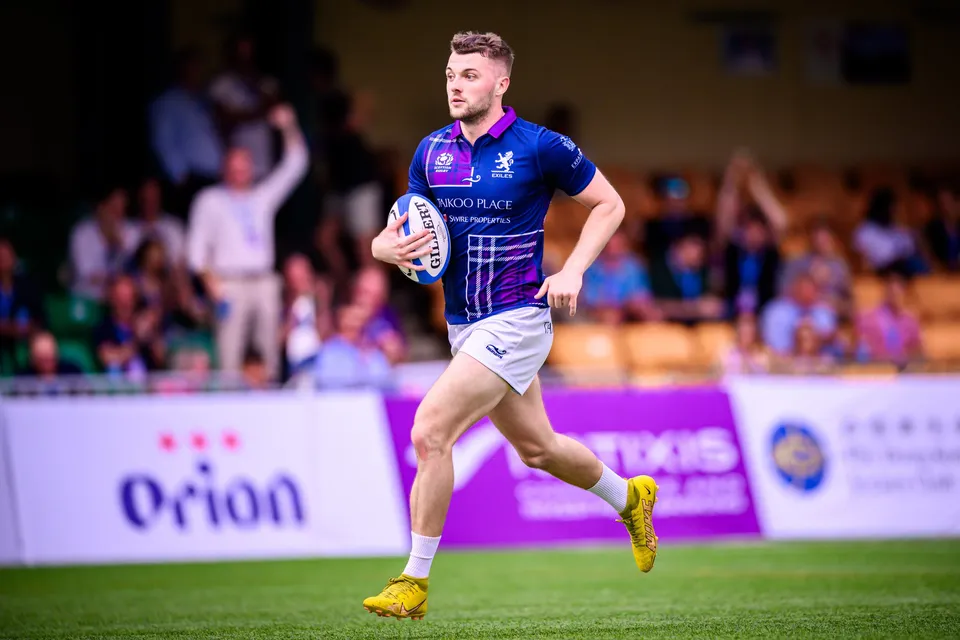 Scottish Exiles player in navy and purple sprints along the touchline with the ball in one hand past Orion boards