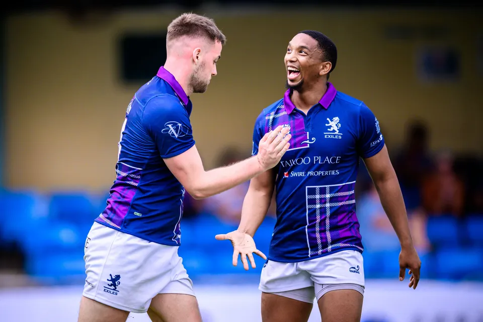 Two Scottish Exiles players in navy and purple laugh and talk, one tapping the other on the chest, blue seats behind