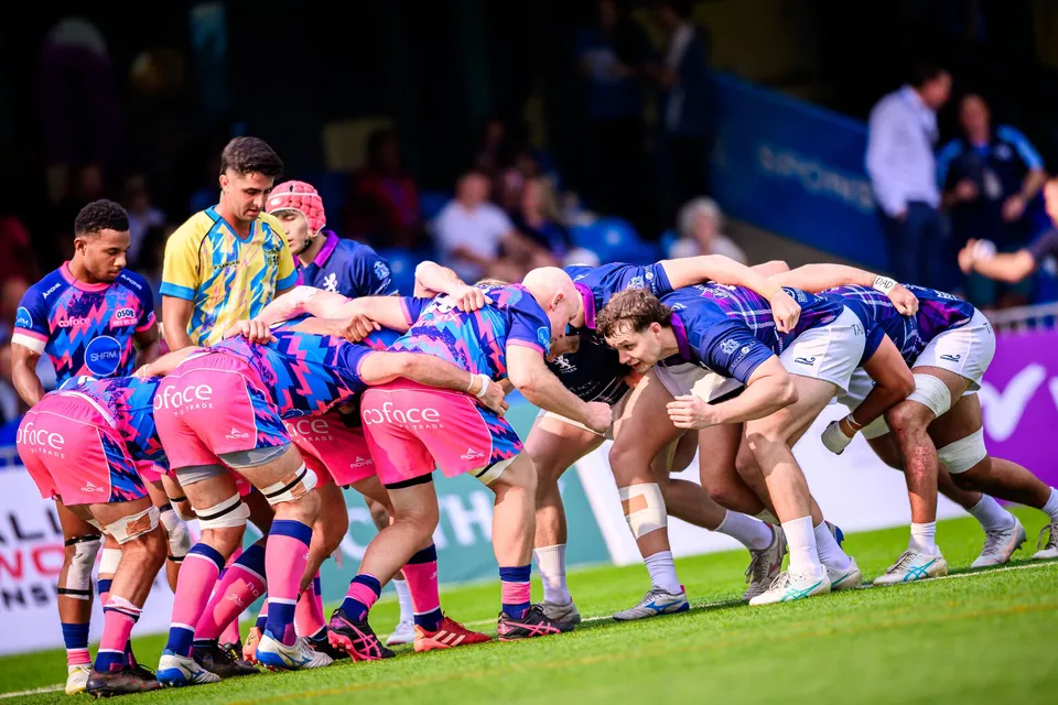 Eight OSOB forwards in pink bind together in a scrum against the Scottish Exiles in navy, on the grass pitch