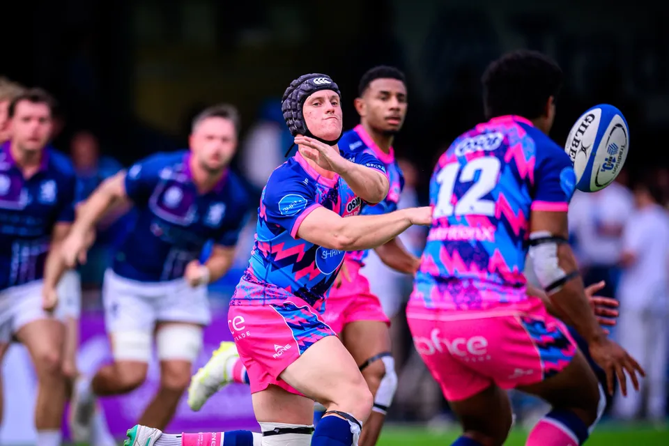 OSOB player in a scrum cap flicks out a pass as number 12 runs across and Exiles defenders in navy close in