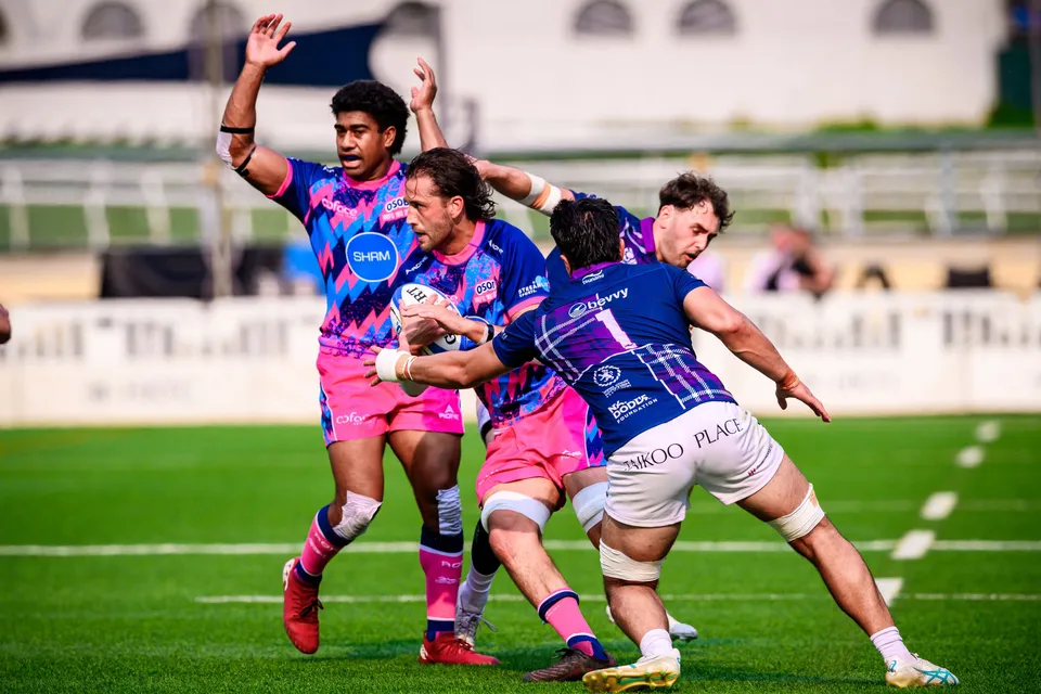 OSOB ball carriers in pink charge into a Scottish Exiles tackler in a Taikoo Place shirt on a bright green pitch