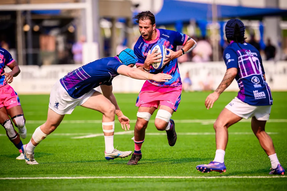OSOB ball carrier clutches the ball in both hands as an Exiles player in a light blue cap tackles his legs
