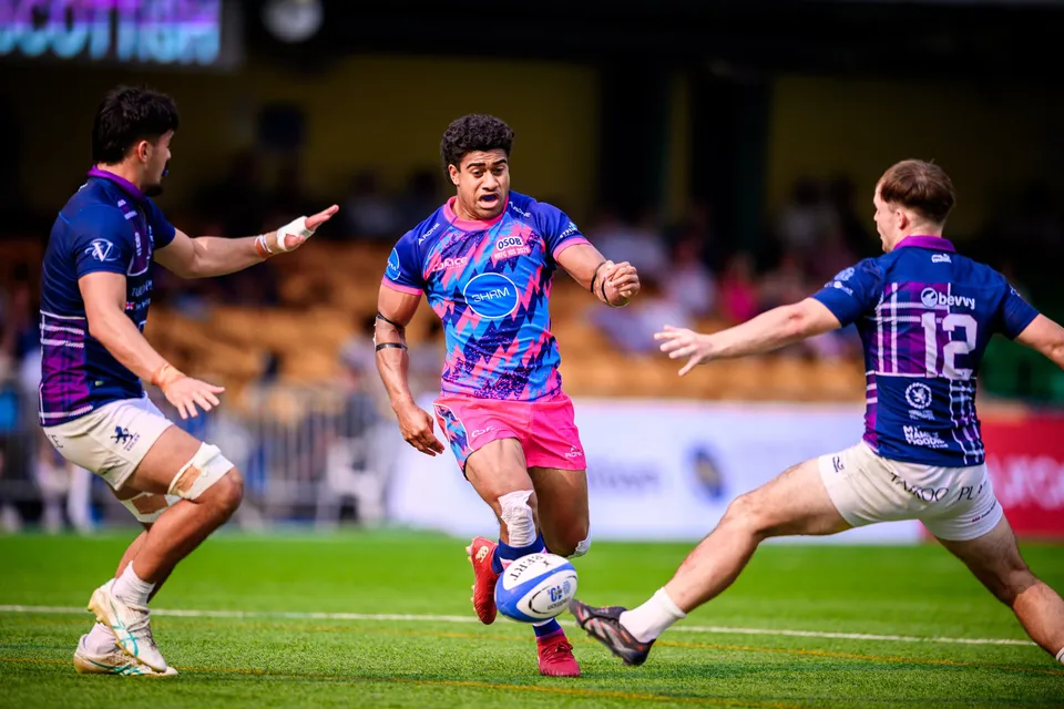 OSOB player in pink chips the ball low between two Scottish Exiles defenders in purple, who stretch out their arms