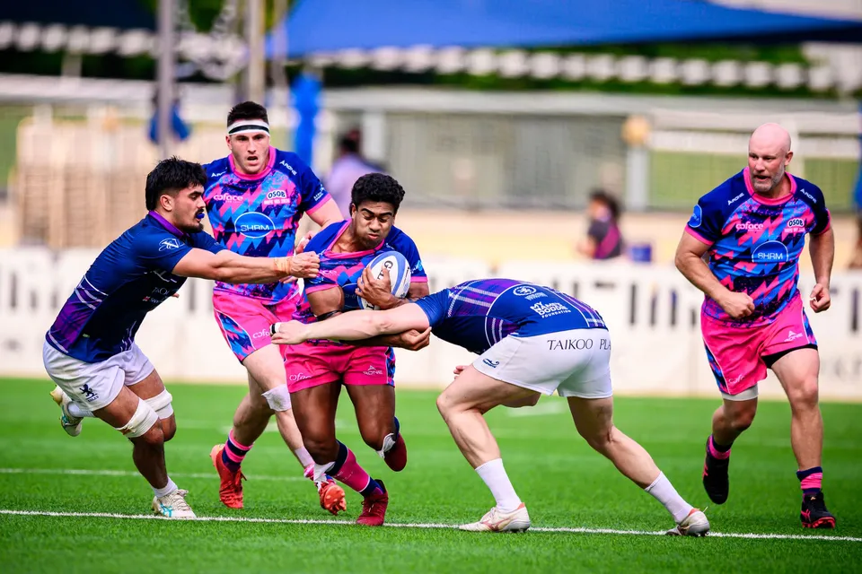 OSOB ball carrier is hit in a low tackle by an Exiles player as teammates in pink, one in a headband, support