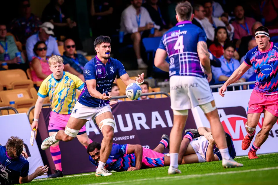 Exiles player in navy passes out of a tackle as a teammate in number 4 towers over a fallen player