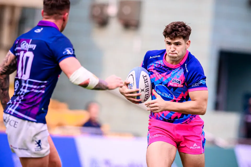 OSOB player in pink holds the ball out in front as an Exiles number 10 in purple closes in, close-up