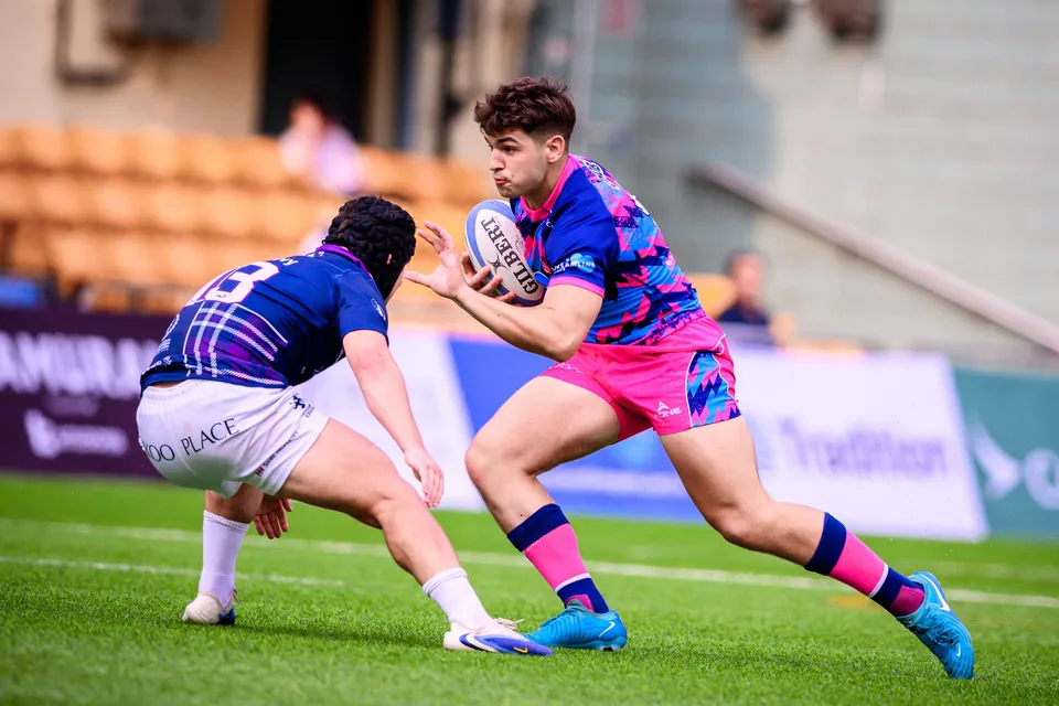 OSOB ball carrier in pink prepares to sidestep a crouching Scottish Exiles defender in a scrum cap on a green pitch