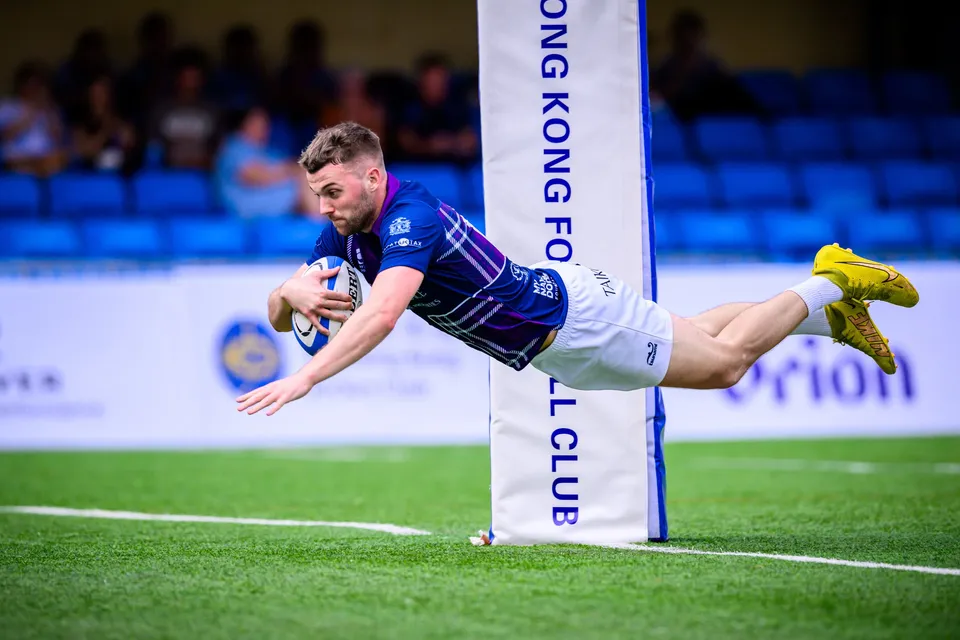 Scottish Exiles player in navy and purple dives full length for the line beside a Hong Kong Football Club post