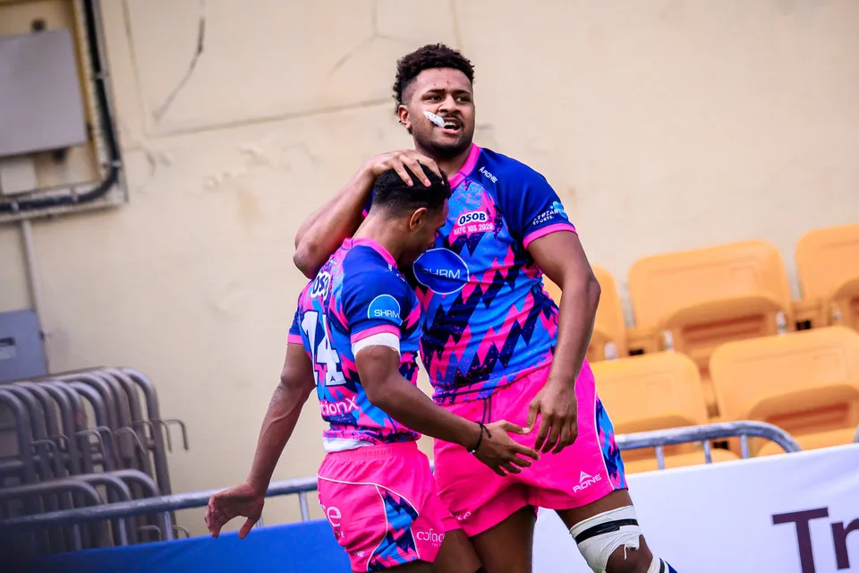 Two OSOB players in pink and blue patterned shirts embrace in celebration in front of yellow stadium seats