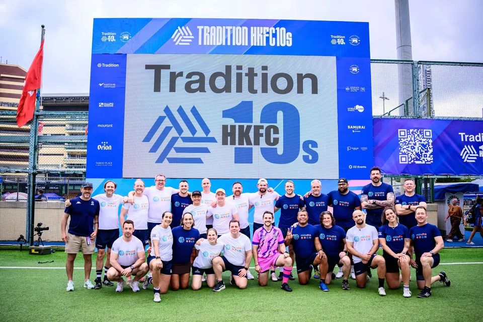 Group of players in white and navy shirts, plus one in pink, pose beneath the Tradition HKFC 10s screen, 2026