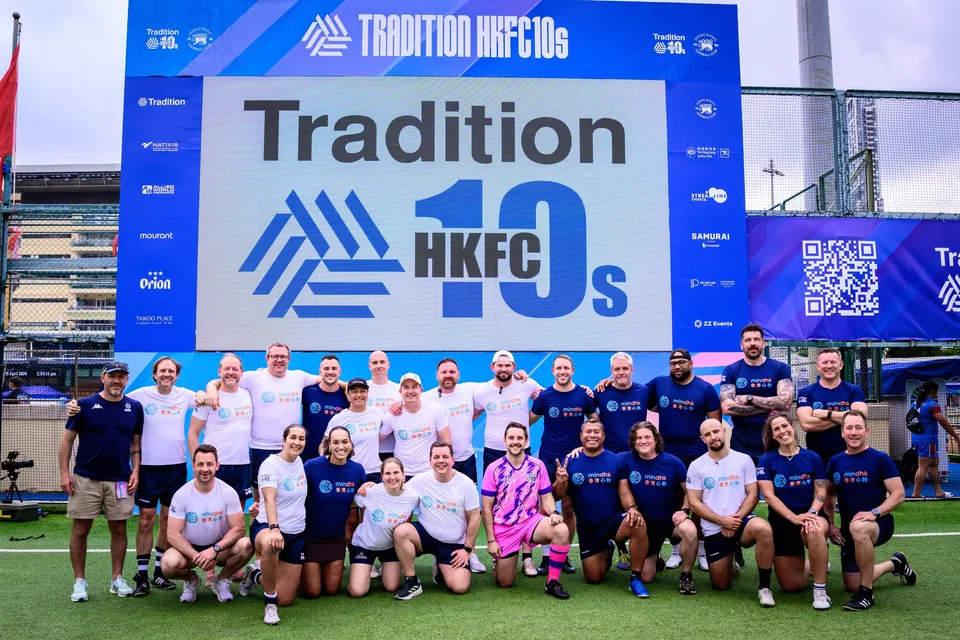 Navy and white shirted squad crouch and stand in front of the Tradition HKFC 10s screen, one player in pink, 2026