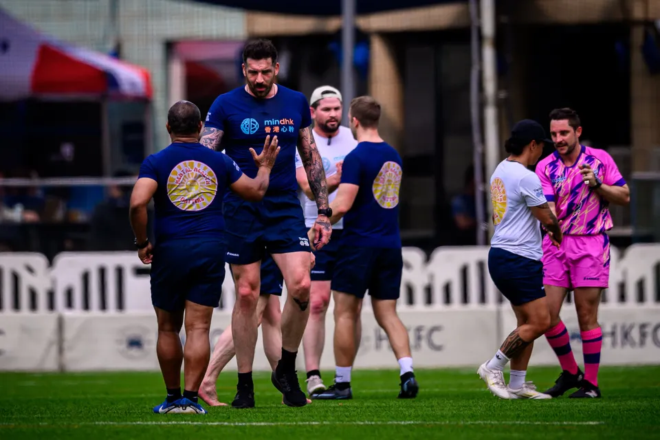 Players in navy shirts walk along the touchline as a man in pink shirt and shorts stands to the right, 2026
