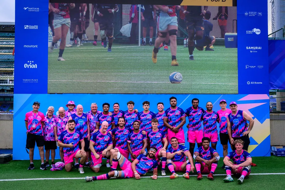 OSOB squad in pink and blue shirts kneel and stand before a screen showing play, with a ball on the grass, 2026