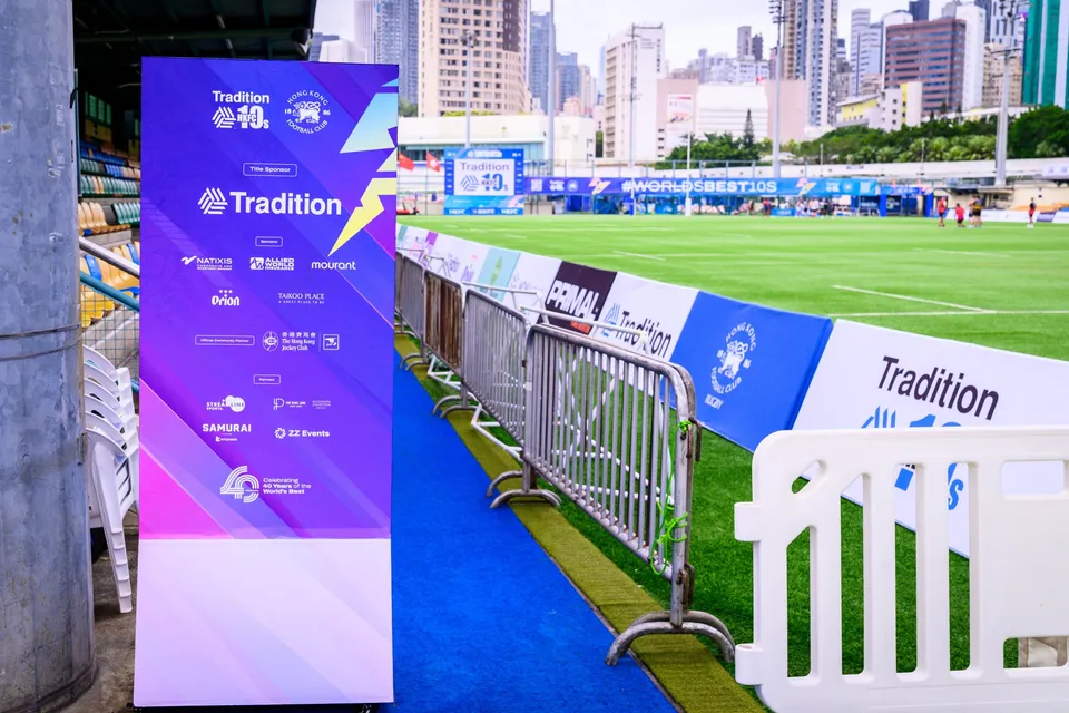 Tradition sponsor board beside a blue walkway and pitchside barriers, with the HKFC pitch behind